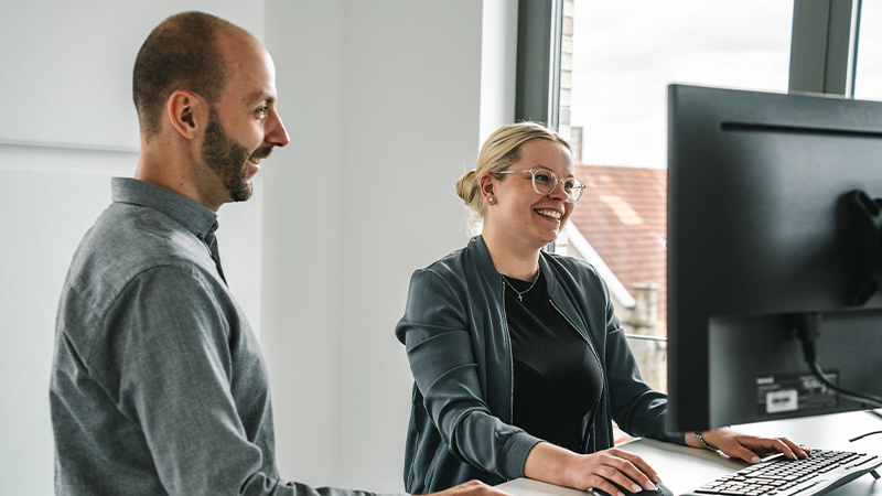 Zwei Mitarbeitende der Steuerkanzlei Runde Glüsing Baune bei der gemeinsamen Arbeit am Stehschreibtisch vor einem Bildschirm